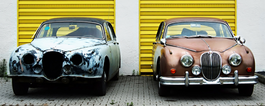Rusting vintage car beside well-kept vintage car outside their lockups